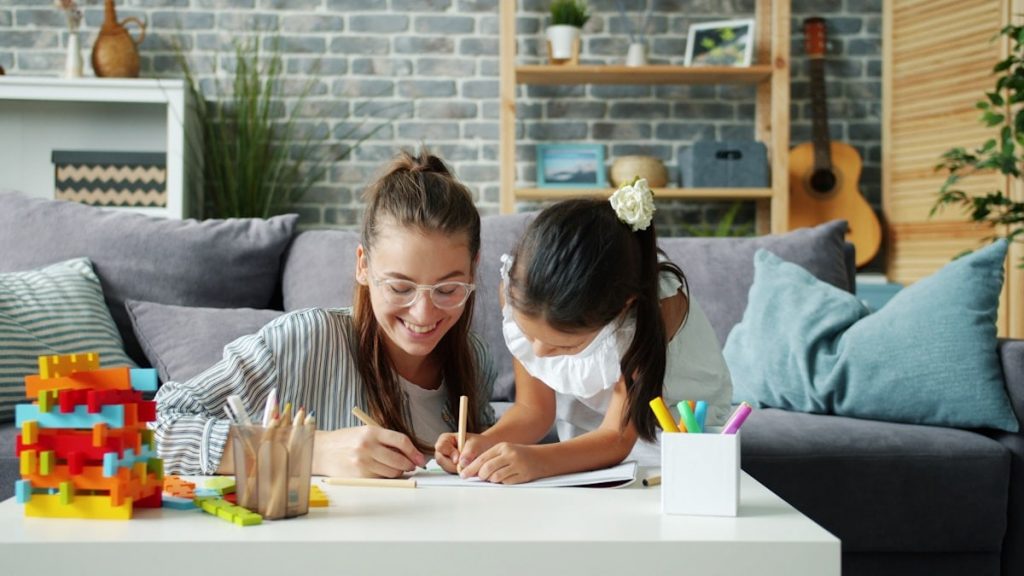 Happy little girl is drawing at home with caring mother using pencils having fun in modern apartment. Creative leisure activity and childhood concept.